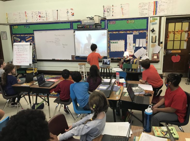 A classroom setting with a student facing another student on the projected screen. 